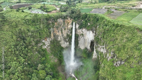Aerial view of Sipiso Piso Waterfall plunging down a steep rocky cliff surrounded by lush green forests and agricultural fields in Merek, North Sumatra, Indonesia.