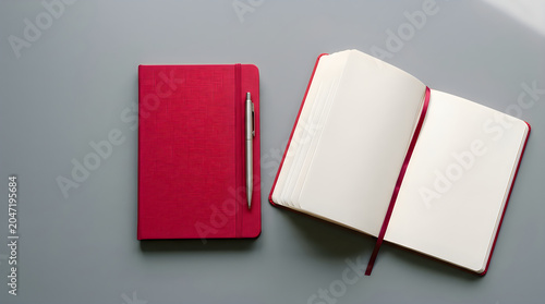 Red notebook rests beside open journal on gray surface. Silver pen tucked into elastic band. Minimalist setup suggests planning, journaling or creative work. Ideal for educational business content