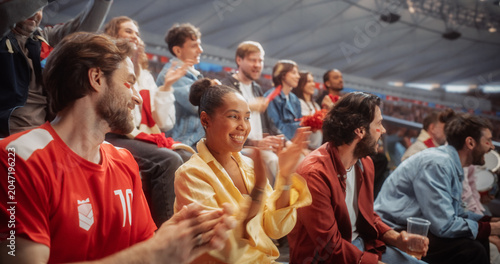 Fotografie Diverse Soccer Football Supporters in Red Sit Side by Side in Packed Stadium Rows, Clapping, Smiling and Cheering While Watching Live Match Play During a Championship Game