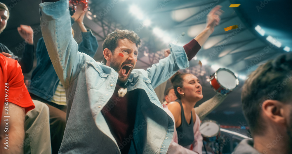 Fototapeta premium Close Portrait of White Male Soccer Football Supporter With Red White Face Paint, Arms Raised Amid Blurred Blue Streamers, Happy Cheer in Packed Championship Stadium Crowd During Cup League Match.