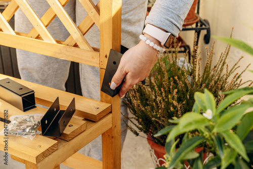 Female hands attaching a black metal bracket to a wooden garden trellis on a balcony, surrounded by potted plants and gardening supplies, conveying DIY, outdoor home improvement and care.