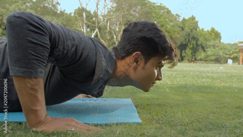 Fit athletic man doing push-ups on a blue exercise mat inside a green park . Closeup shot of a young Indian male practicing daily workout session in the morning - healthy lifestyle