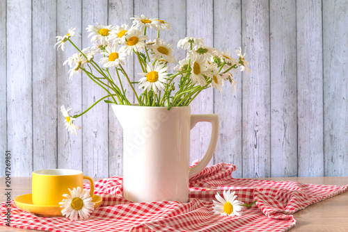 Summer season still life; yellow cup of tea and bouquet of white daisy flowers on a red and white checkered napkin against wooden background