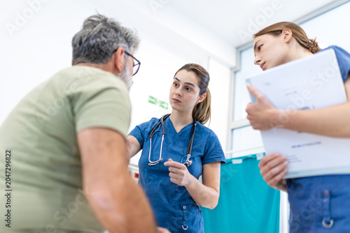 Female medical doctors consulting senior male patient in clinic, healthcare concept. Two professional nurses listening to elderly man case history during clinical examination in hospital ward room.