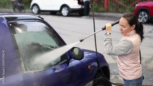 Watch as a woman meticulously washes her blue car at a self-service car wash. The sunlight glints off the water, highlighting the vibrant color and the joy of a clean vehicle. Slow motion 4k footage.
