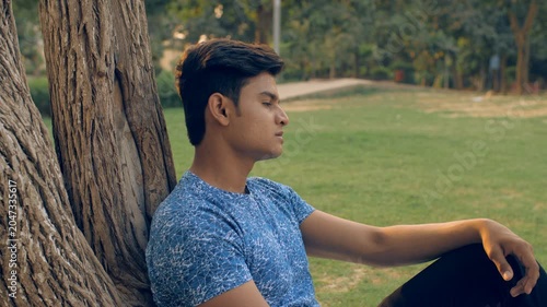 Upset young man thinking about his life while sitting near a tree trunk in the park. A concerned youngster wearing a printed T-shirt feeling lonely and sad while sitting on the garden ground - anxi...