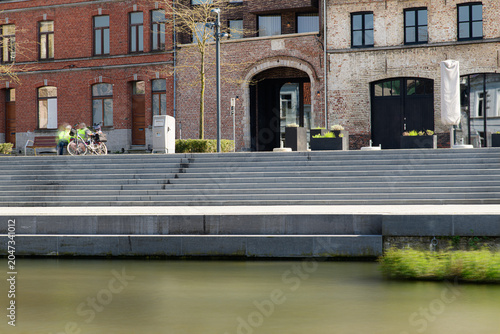 An urban waterfront scene with wide concrete steps descending to a calm canal, parked bicycles by a bench, red brick buildings and soft spring sunlight conveying a peaceful city leisure mood.