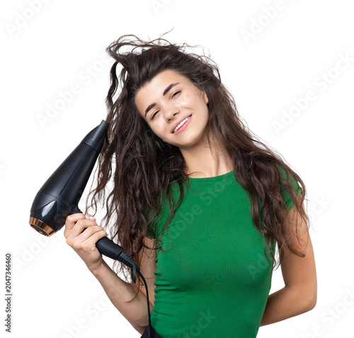 A cute young girl dressed in a green top dries her beautiful long silky hair with a hair dryer against a white background.