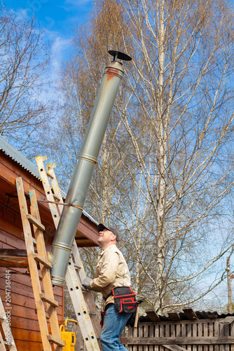 A man on a ladder inspecting a high metal chimney pipe of a wooden country house. Checking the heating system and home maintenance concept.