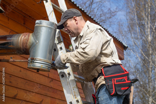 A worker on a ladder tightening fasteners on a metal chimney tee pipe near a wooden house wall. Final stage of heating system installation.