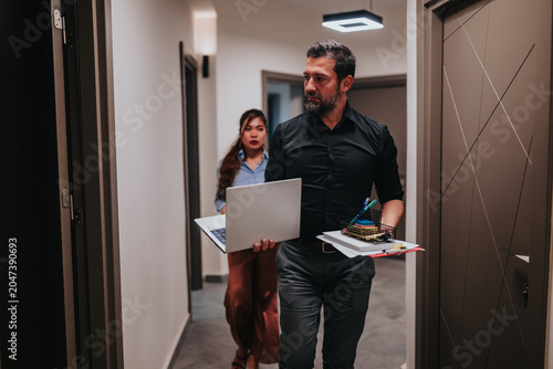 Cuadro en lienzo A male and female business teammates walk down an office hallway carrying a laptop and documents