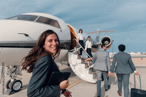 Professional young businesswoman in navy blazer holding digital tablet greets arriving business executives with warm smile at white private corporate jet aircraft on sunny coastal airport tarmac with