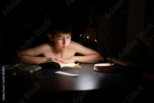 Photography A preteen boy reads a book by a warm desk lamp in dark room, absorbed and calm as he studies or reads for pleasure late at night