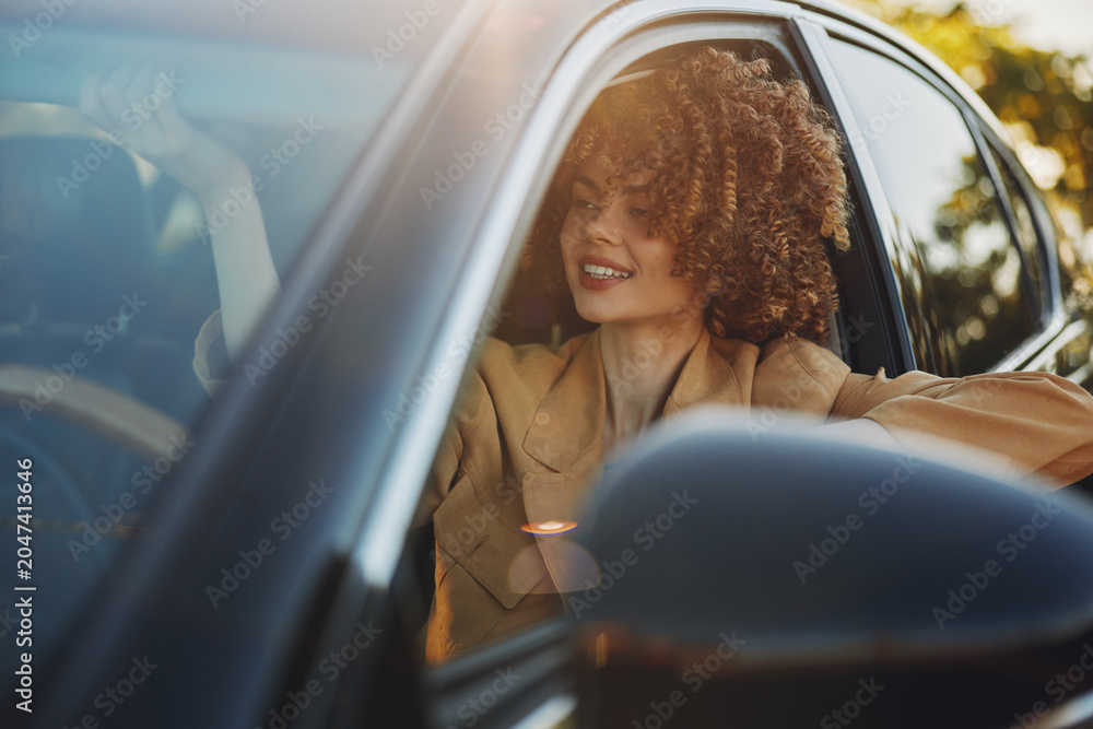 Fototapeta premium Smiling woman with curly hair sits in a car, sunlight glints on the window, warm retro tones create a relaxed lifestyle mood and sense of casual travel