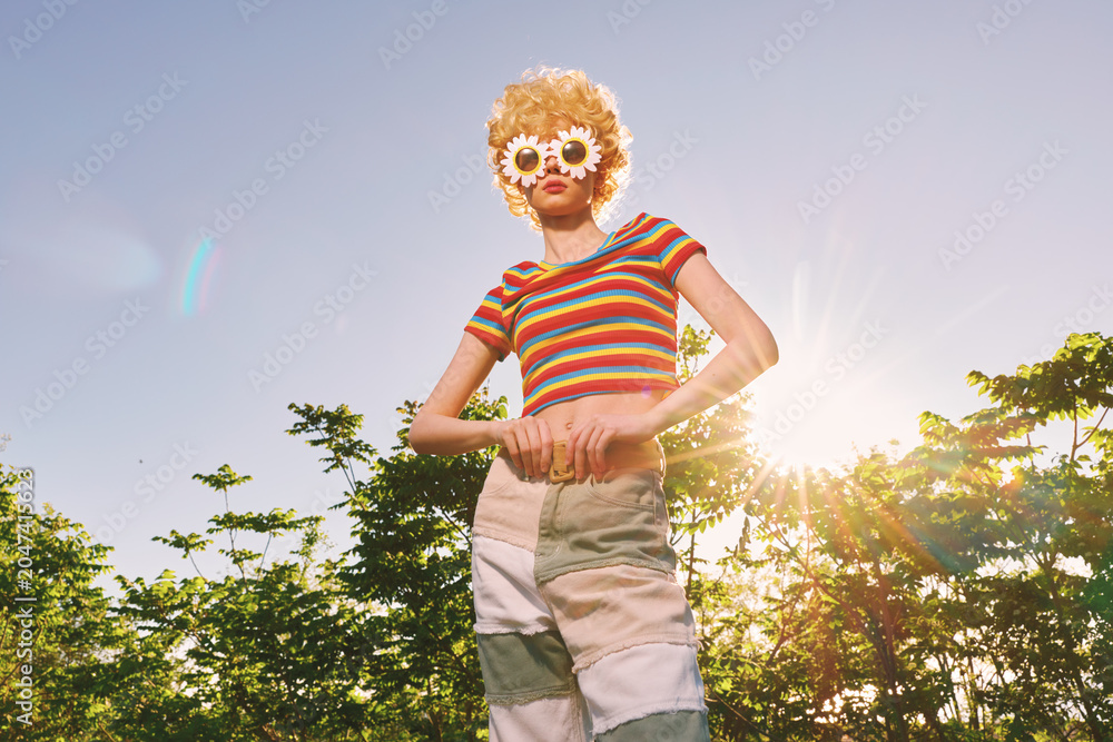 Naklejka premium A stylish woman poses outdoors in a sunny park, wearing a striped tee and round floral sunglasses. Retro film color tones evoke carefree summer vibes and playful fashion.