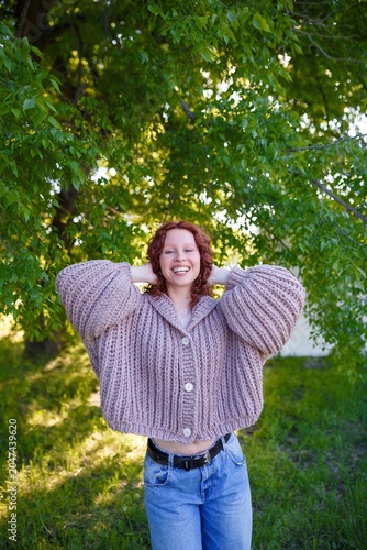 Redhead teenage girl model in chunky knit sweater and jeans on green park background. Bulky knitwear, clothing and style concept