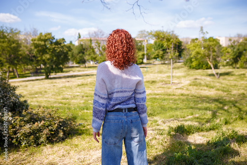 Young woman with curly red hair stands back to camera on green natural background. Wearing jeans and knit blue striped sweater