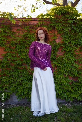 Redhead teenager girl model in knitted purple transparent sweater and white silk skirt posing outdoors. Clothing and fashion concept