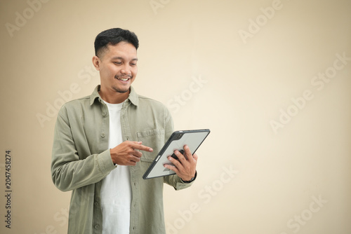 Young Asian man tapping on a digital tablet screen while smiling, using the device in casual olive green shirt against beige studio background