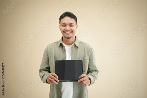 Happy young Asian man showing a digital tablet with blank screen facing camera, presenting the device with both hands in casual studio portrait