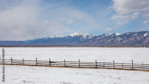 Wide winter landscape of snowy steppe with a rustic wooden fence and distant snowcapped mountain range under cloudy sky in Buryatia, Siberia, Russia