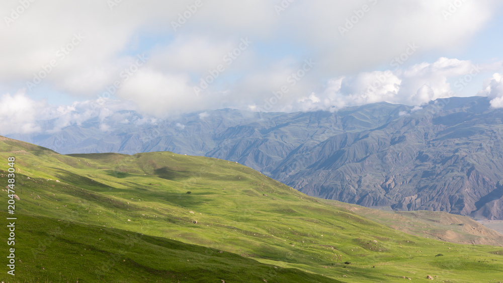 Fototapeta premium Scenic trekking road leading toward Mount Shalbuzdag, with rolling green alpine hills and dramatic Caucasus mountain ridges under cloudy sky, Dagestan, Russia