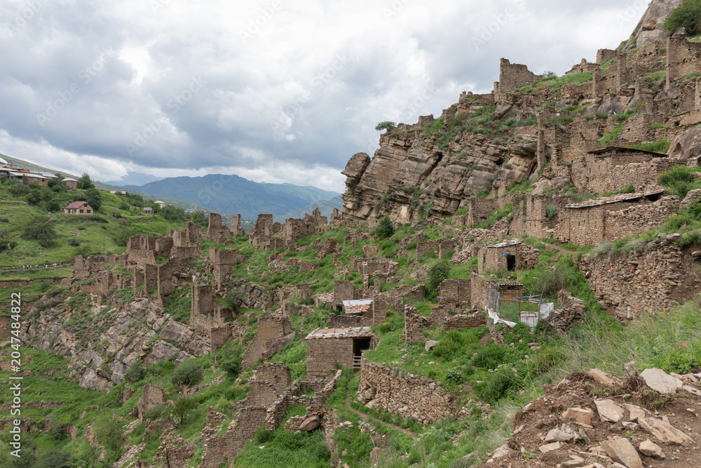 Fototapeta premium Wide view of abandoned stone houses and fortress ruins on a steep green mountainside in Kahib, Dagestan Republic, Russia, under dramatic cloudy sky