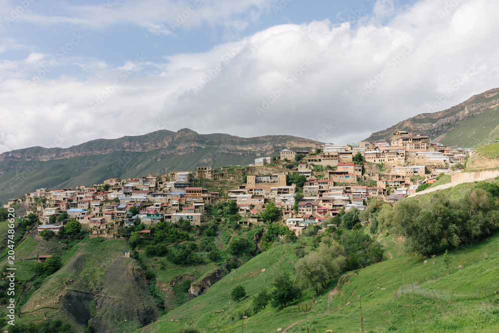 Obraz premium Wide landscape view of Chokh aul, a traditional mountain village built on a steep hillside in Dagestan, Russia, with terraced homes, green slopes and cloudy sky
