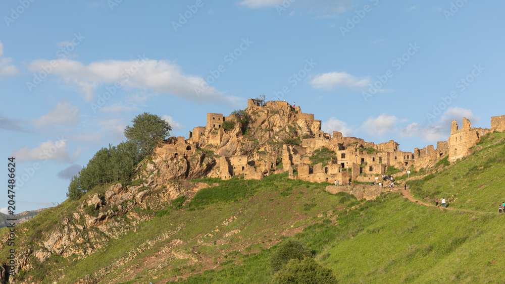 Fototapeta premium Panoramic view of the ruins of Gamsutl, an abandoned stone mountain village on a hillside in the Republic of Dagestan, Russia, under blue sky and clouds