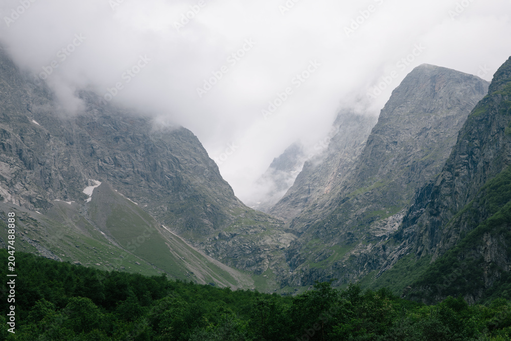 Obraz premium Dramatic view of Midagrabin Waterfalls gorge with steep rocky mountains, low clouds and green forest in North Ossetia–Alania, Russia, Caucasus region