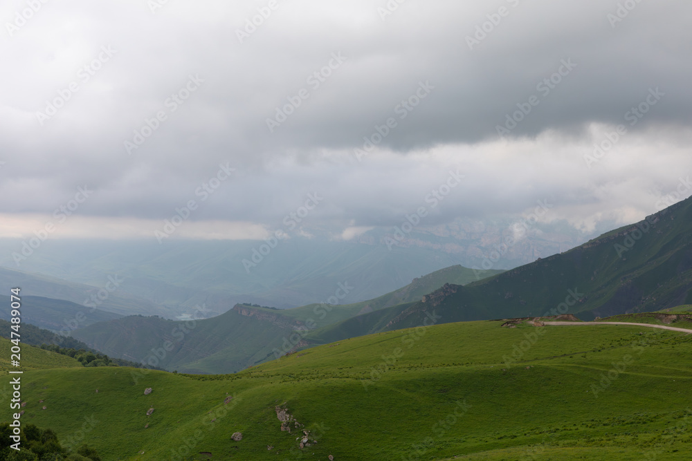 Fototapeta premium Scenic view of rolling green alpine hills and distant mountain ridges under dramatic storm clouds near Aktoprak, Kabardino-Balkaria, Russia