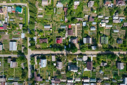 Top down aerial view of a suburban neighborhood with small houses, garden plots, greenhouses, and dirt roads surrounded by lush summer greenery