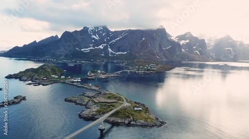 A bridge connects islands in Norway as boats move slowly in the water. Clouds float above and hills rise in the distance. This view shows the landscape and water.