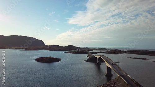 A bridge connects islands in Norway as boats move slowly in the water. Clouds float above and hills rise in the distance. This view shows the landscape and water.