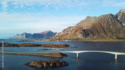 A bridge connects islands in Norway as boats move slowly in the water. Clouds float above and hills rise in the distance. This view shows the landscape and water.