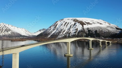 A bridge connects islands in Norway as boats move slowly in the water. Clouds float above and hills rise in the distance. This view shows the landscape and water.