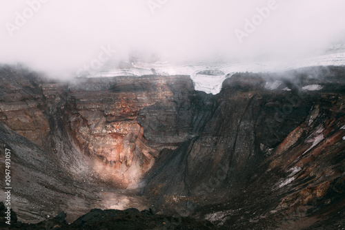 Dramatic view into the Tolbachik volcano crater with layered lava rock, snow patches and low clouds. Remote wilderness scenery in Kamchatka Krai, Russia