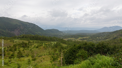 Wide scenic view of the Vachkazhets mountain range with lush green valley, forested hills and distant peaks under overcast clouds in Kamchatka Krai, Russia