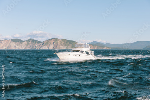 Canvas Print White motor yacht speeding through choppy ocean waves with rocky coastal mountai