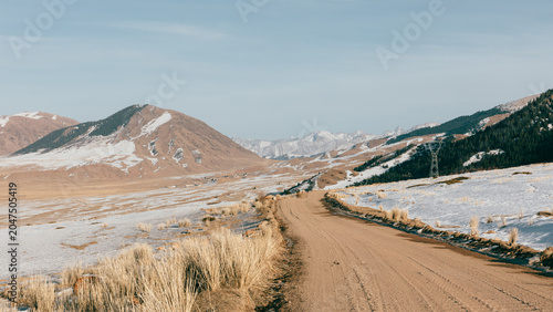 Scenic winding dirt road crossing a snowy mountain valley with dry grass and distant peaks, rural highlands of Kyrgyzstan in Central Asia, winter travel concept