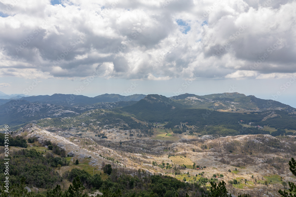 Fototapeta premium Wide panoramic view of rocky mountains, forests and valleys under dramatic cloudy sky in Lovcen National Park, Montenegro, a scenic nature travel destination