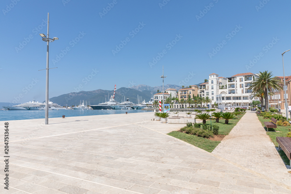 Fototapeta premium Tivat, Montenegro - August 11, 2025: Sunny waterfront view of Porto Montenegro marina with luxury yachts, modern promenade, palm trees and coastal buildings in Tivat, Montenegro, on a clear summer day