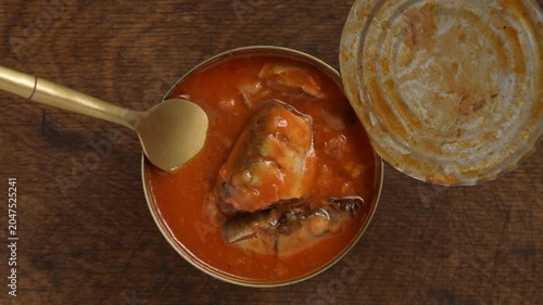 Top view of opened canned fish sprats in red tomato sauce with a golden fork on a dark wooden table, close up