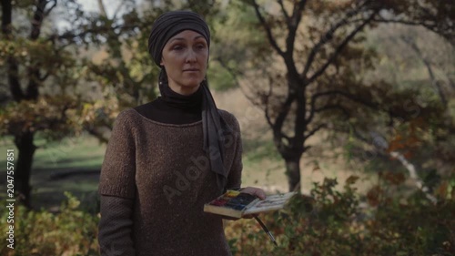 Artist with watercolor palette in hand looking thoughtfully in front of her in autumn forest