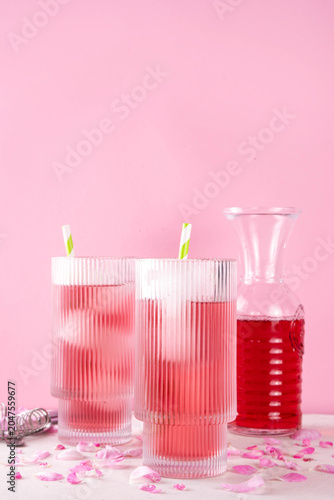 Rose petals iced tea, tasty infused water or lemonade with organic rose syrup, in two tall glasses, with ice cube and rose petals on white pink background