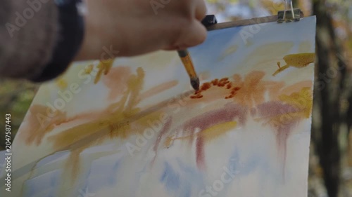 Close-up, the artist in front of the easel paints a landscape of an autumn forest