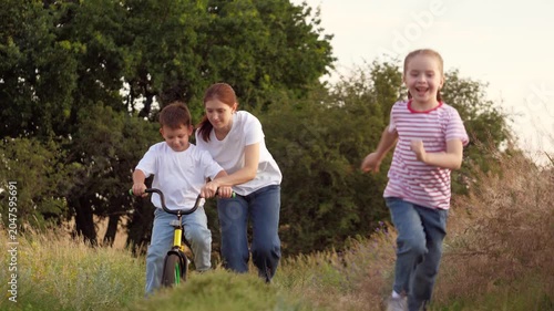 Smiling girl runs while mother teaches little boy to ride bicycle on grassy rural road at sunset. Mother with girl and boy practice bicycle riding in field. Caring mother with boy and girl on vacation