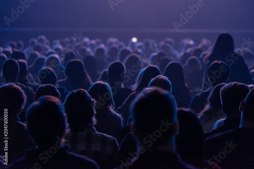 Large seated audience watching stage performance in darkened venue, blue lighting creates moody atmosphere.