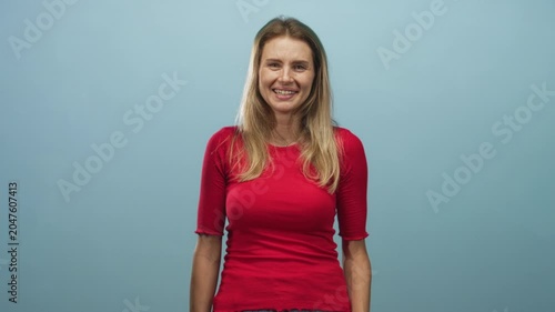 Woman blonde and young smiling and laughing with hand covering face, wearing red top and jeans while standing against a pale blue studio backdrop; joy playfulness.