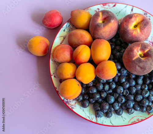 Ripe apricots, flat peaches and blueberries arranged on a decorative plate with purple background and copy space. Fresh summer fruit concept for healthy eating, dessert, harvest and nutrition.